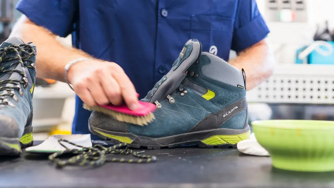 A person is cleaning a hiking boot with a brush, demonstrating how to maintain shoes.