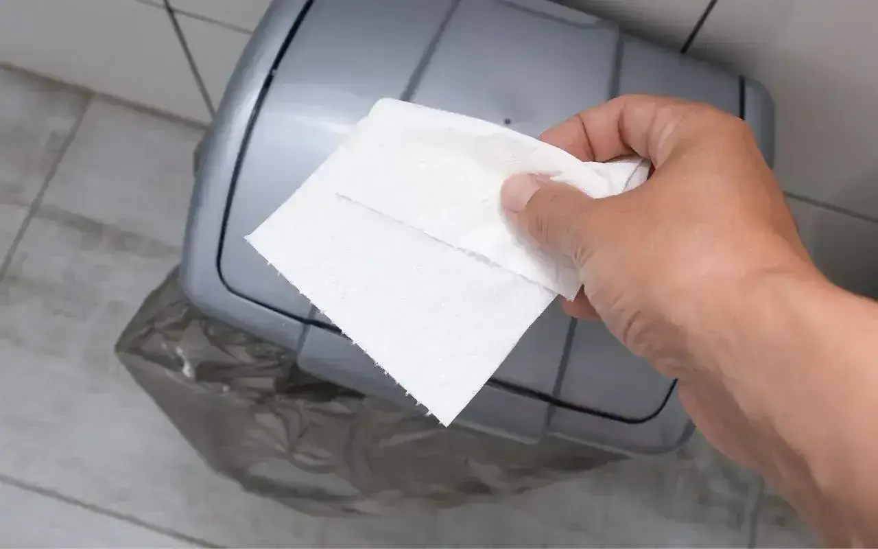 Dłoń z ręcznikiem papierowym zbliża się do kosza. Właśnie tu, gdzie wyrzucać ręczniki papierowe, by zachować czystość.