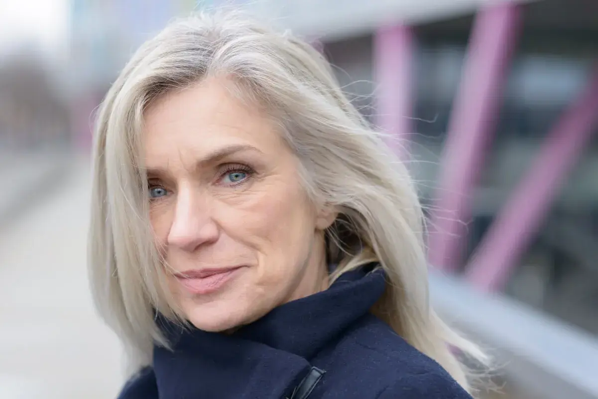 Donna con capelli biondo cenere, un sorriso discreto e occhi azzurri. Nonostante i capelli bianchi, il suo look è curato, quasi a voler camuffare capelli bianchi con un'eleganza naturale.
