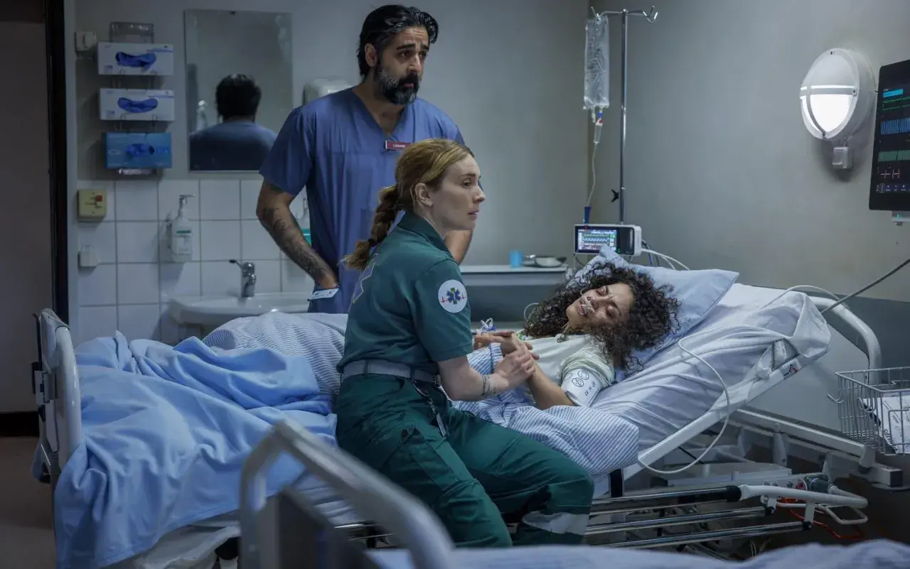 En ambulanssjuksköterska håller en patients hand medan en läkare ser på. Scen från en vaka serie.