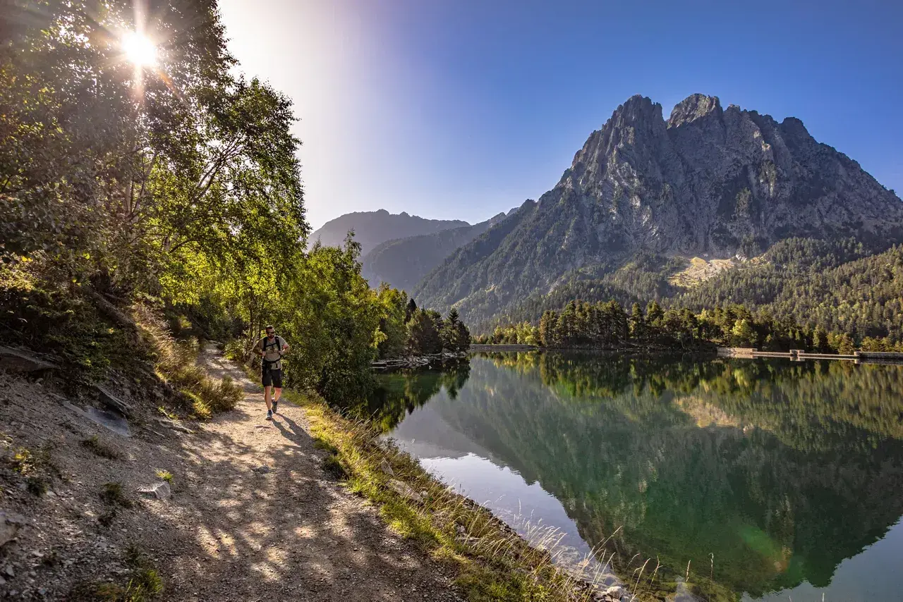 Senderista camina por el sendero junto al estany de Basturs, reflejando la majestuosa montaña y el bosque en sus aguas tranquilas.