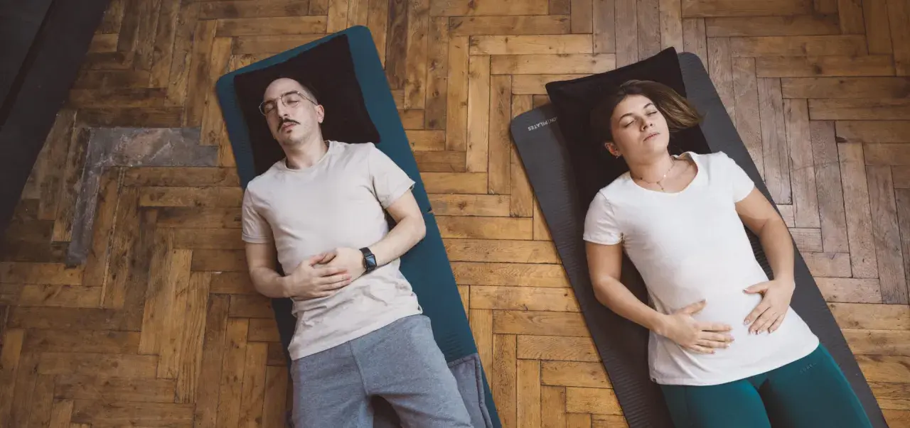 Un couple pratique la respiration profonde technique sur des tapis de yoga. Ils sont allongés sur le dos, les yeux fermés, les mains sur le ventre.