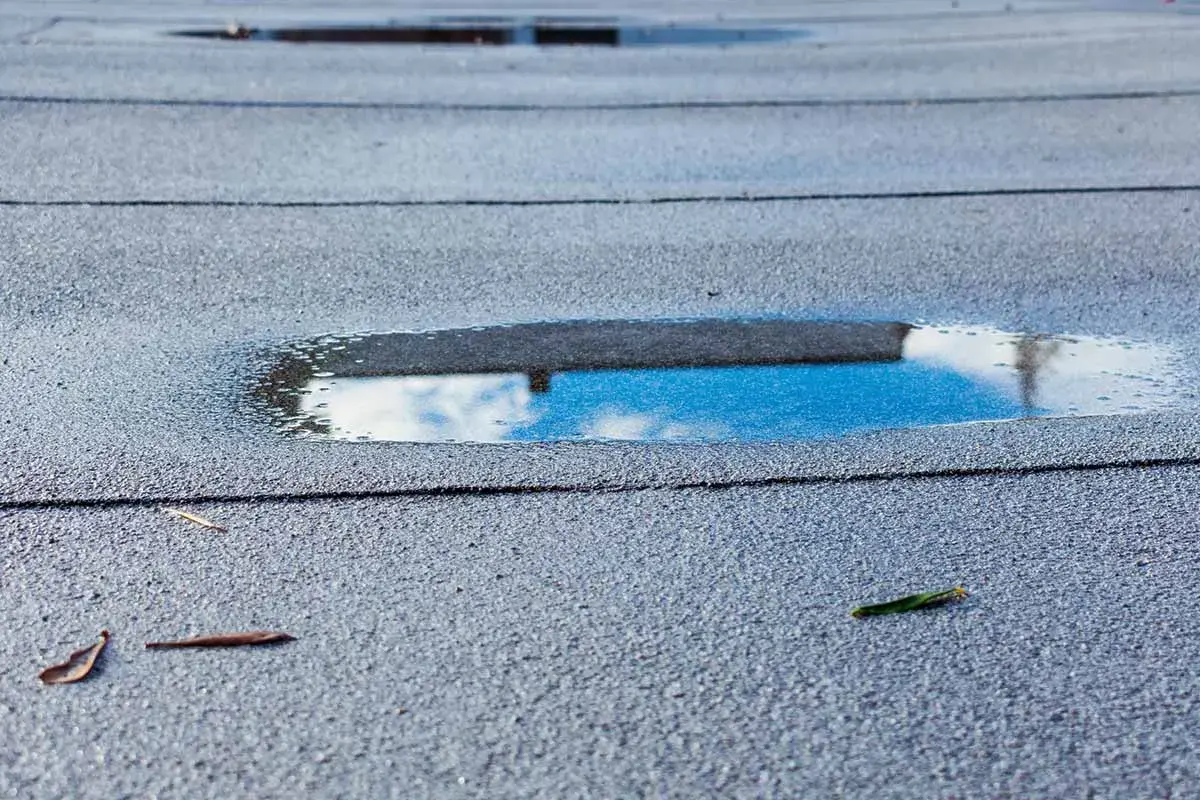 Pfützen auf der Dachpappe spiegeln den Himmel. Feuchtigkeit unter Dachpappe deutet auf ein Problem hin.