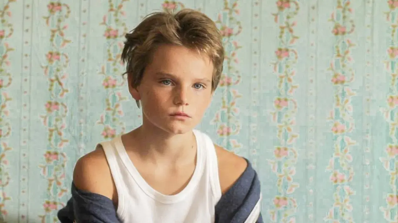 Un niño con cabello rubio y ojos azules, con una camiseta blanca y una chaqueta azul, parece un tomboy que es.
