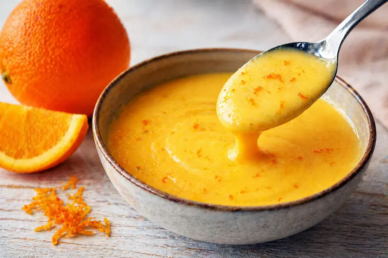 Une cuillère dégouline d'une sauce maltaise onctueuse, parsemée de zestes d'orange. Une orange entière et une tranche sont à proximité.