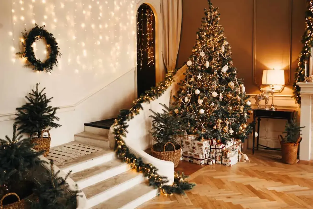 Un albero di Natale scintillante, decorato con nastri natalizi per albero, addobbi dorati e luci calde. La scala è adornata da ghirlande luminose e piccoli abeti in cesti.