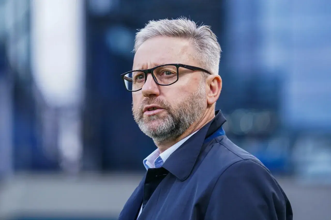 Mężczyzna w okularach, z siwą brodą i włosami, ubrany w ciemną kurtkę. Wygląda jak trener u21, skupiony na boisku.