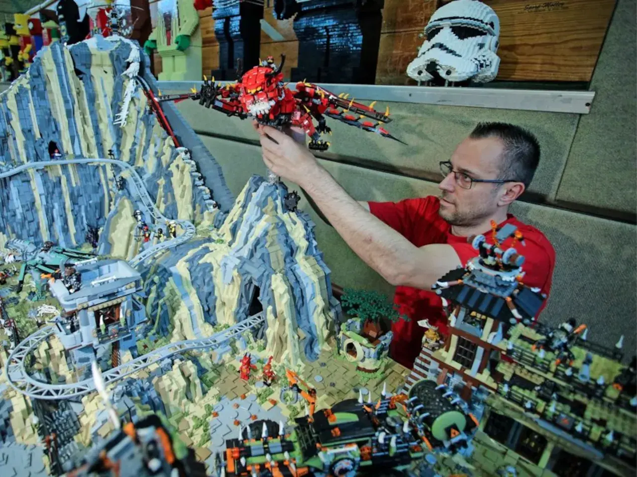 Mann mit Brille platziert roten Drachen in einem riesigen Lego-Set mit den meisten Teilen, das eine Berglandschaft mit Achterbahn und Gebäuden zeigt.