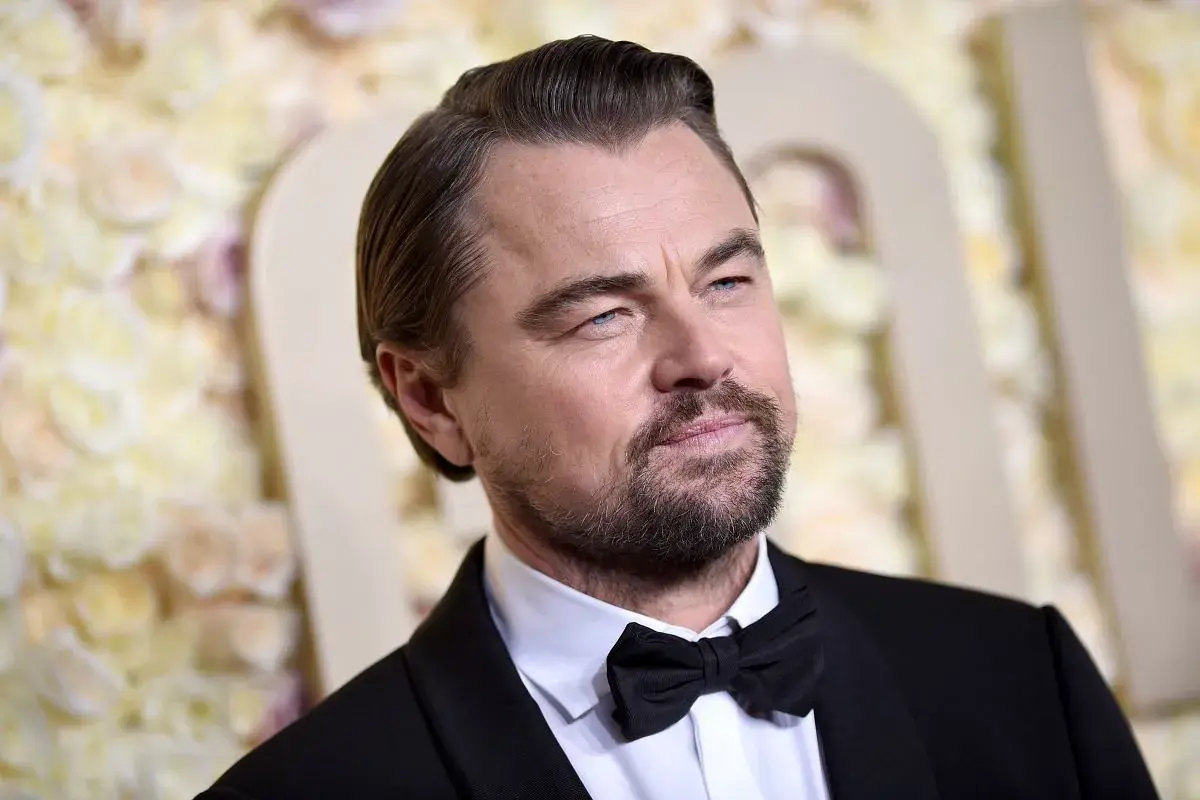 Leonardo DiCaprio wiek: aktor w czarnym smokingu z muszką, z siwiejącymi włosami i zarostem, na tle kwiatowej dekoracji.