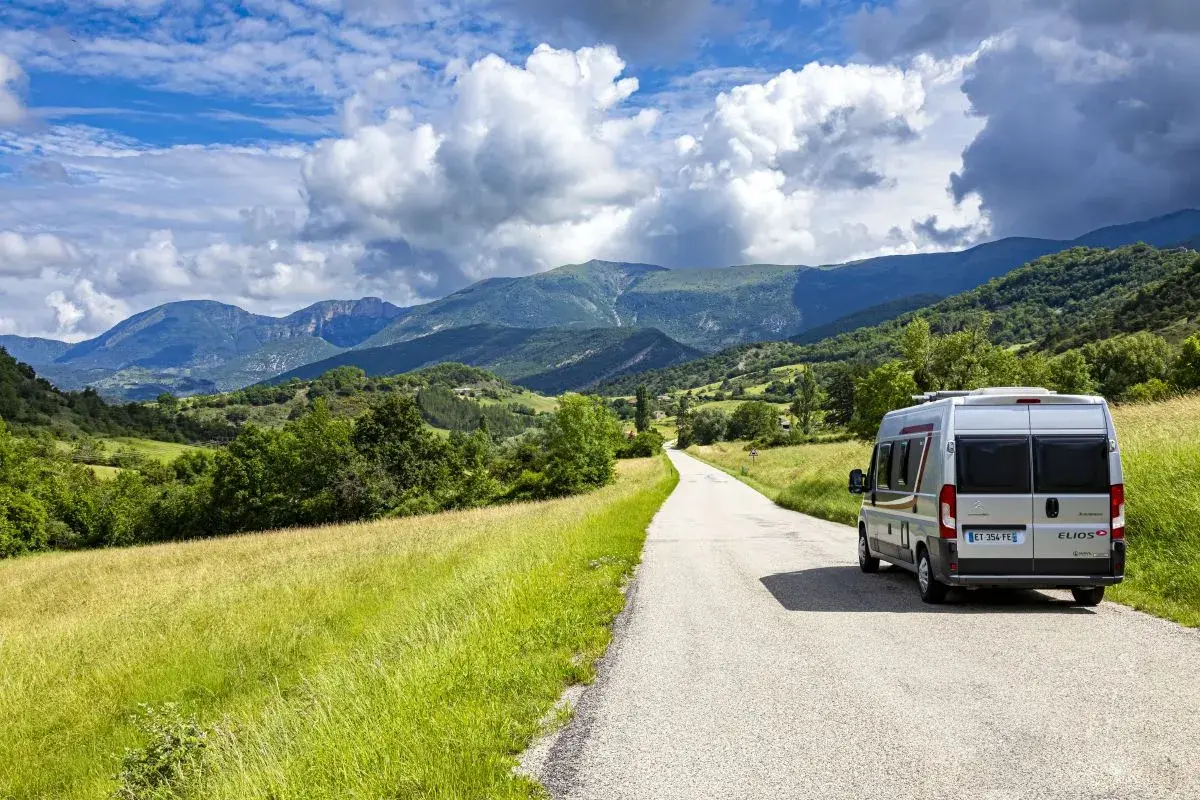 Un camping-car Elios sillonne une route sinueuse à travers les paysages verdoyants du Vercors, sous un ciel bleu parsemé de nuages.