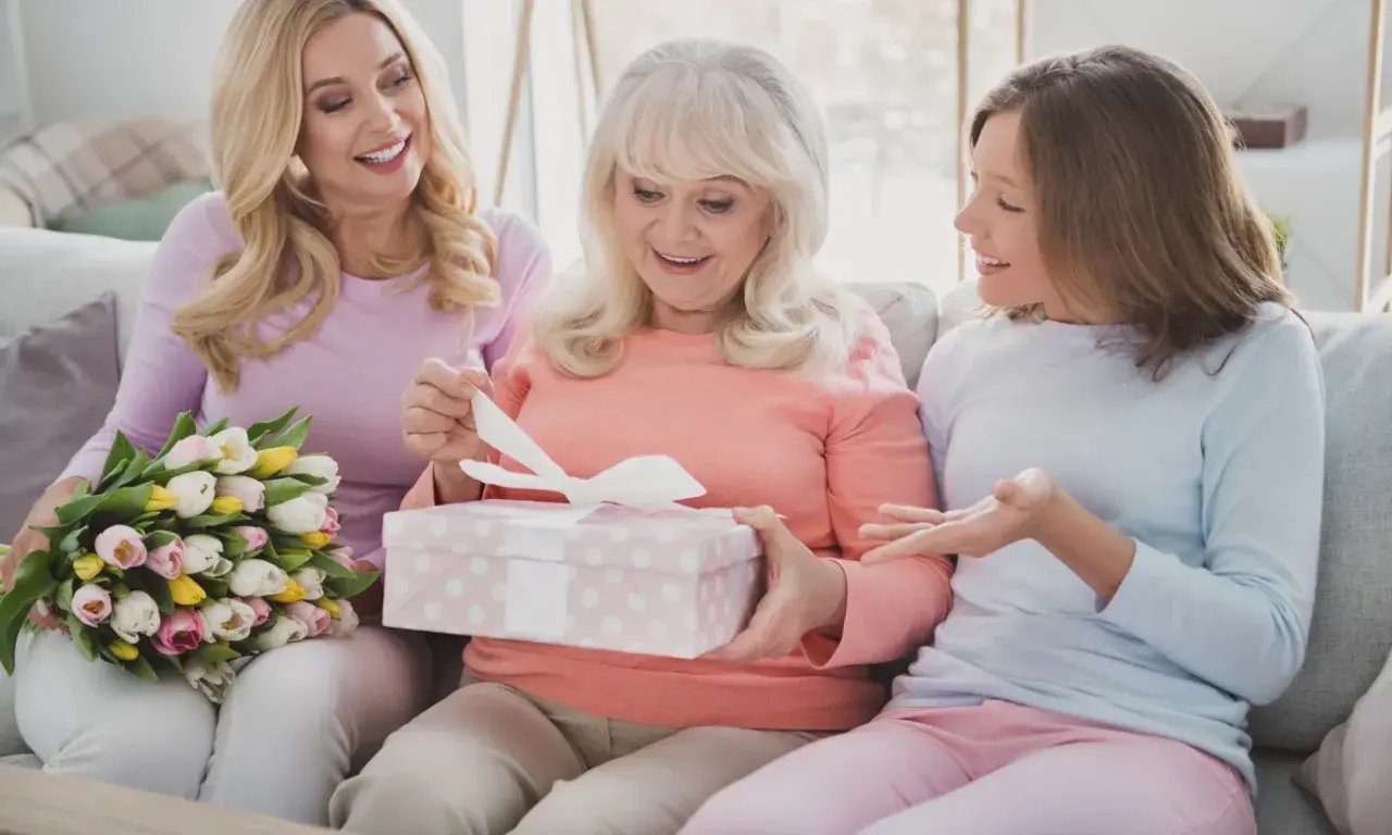 Trzy kobiety na kanapie: starsza otwiera prezent, młodsza trzyma bukiet, najmłodsza gestykuluje.