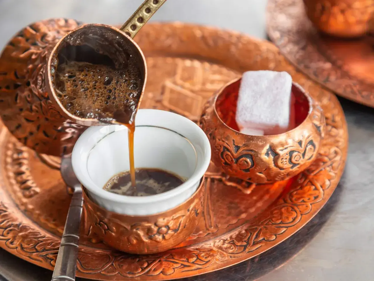 Frisch aufgebrühter, bester türkischer Kaffee fließt aus einer Kupferkanne in eine kleine Tasse. Daneben liegen süße Loukoum-Würfel.