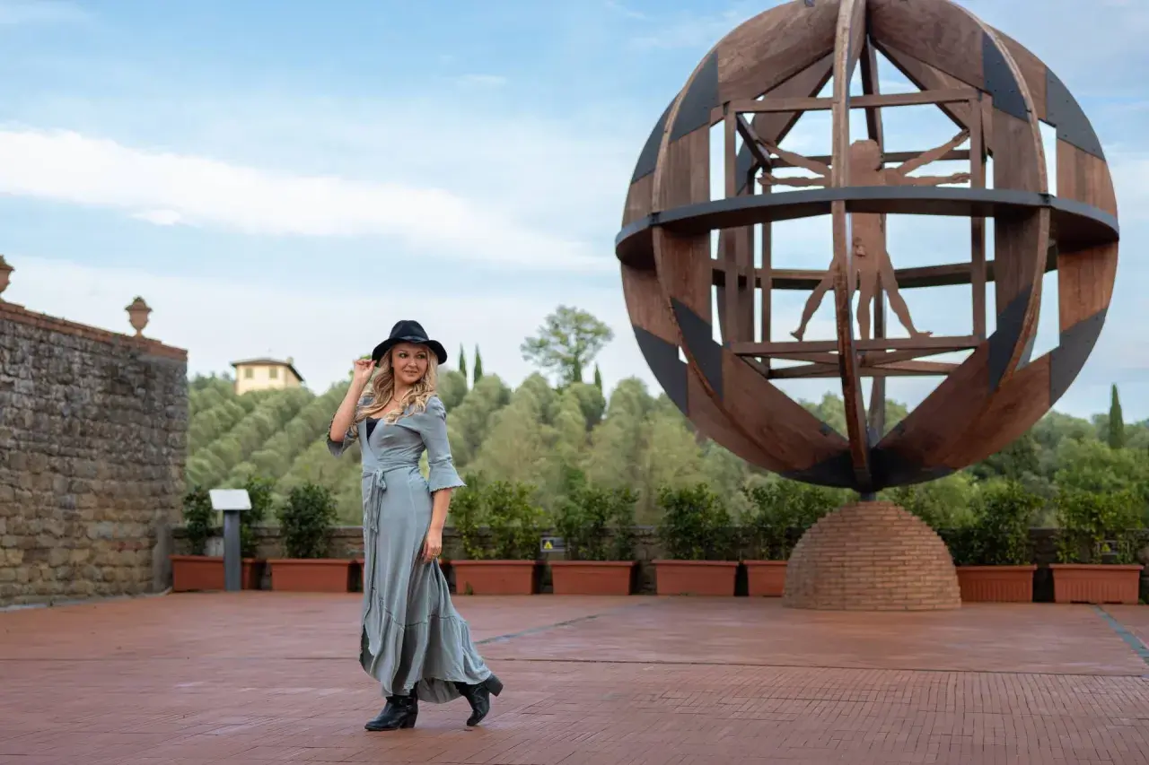 Donna con cappello nero e abito lungo in un'area panoramica del vinci centro storico, con la scultura dell'Uomo Vitruviano sullo sfondo.
