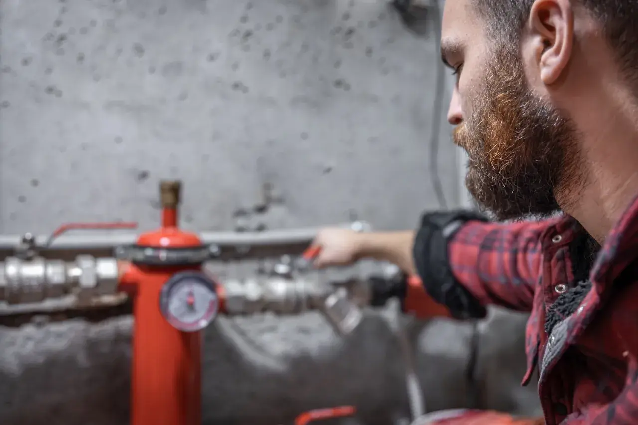 Uomo con barba controlla un manometro su un tubo rosso. Sento rumore nei tubi dell'acqua, forse un problema da risolvere.