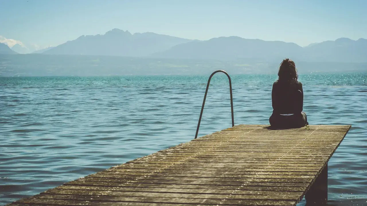 Mujer sentada en un muelle, contemplando el lago y las montañas. Pensamientos de frases de preocuparse por alguien flotan en el aire.