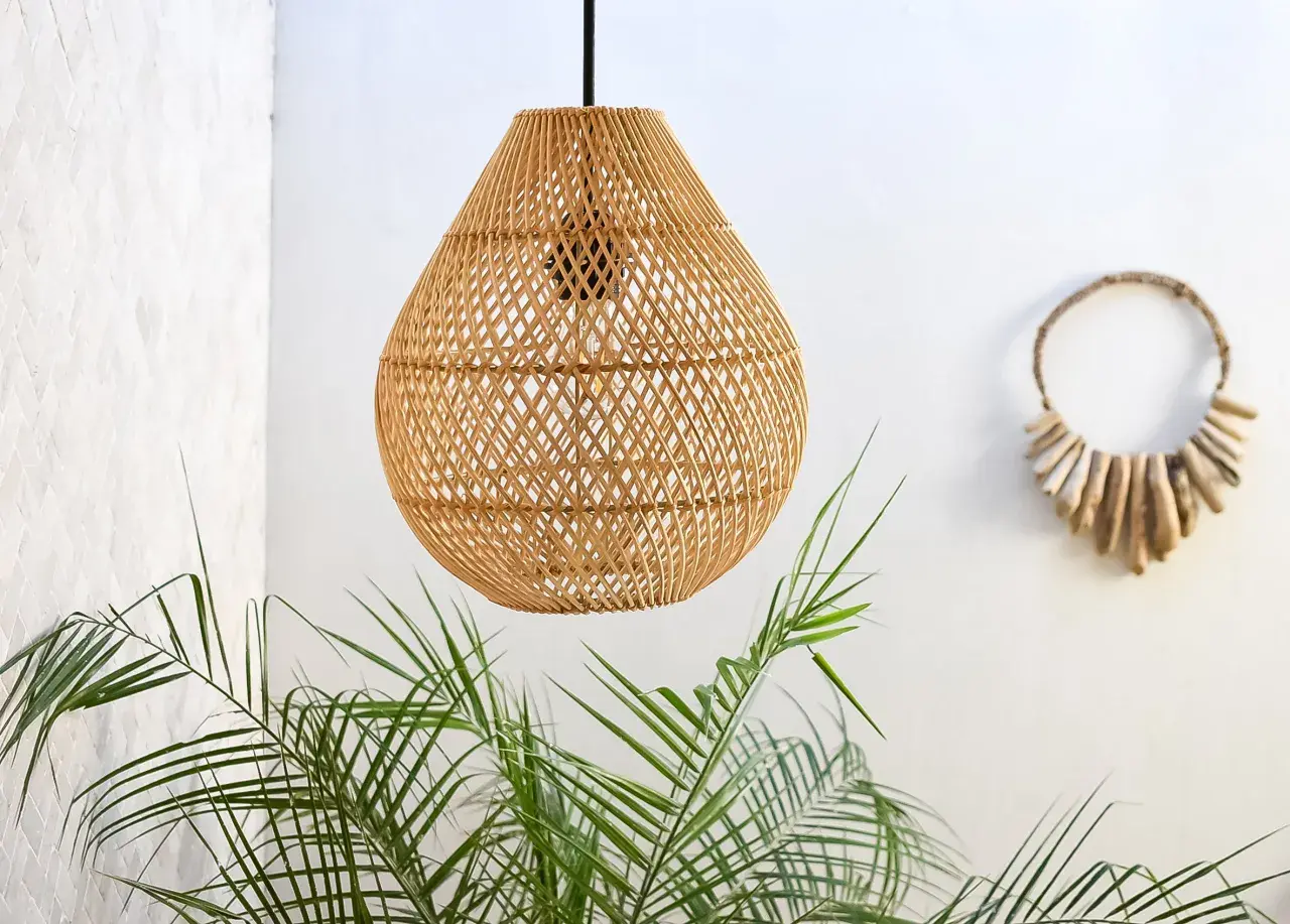 Lámpara colgante de ratán con un diseño de tejido abierto, como un adaptador de pantalla para lámpara, junto a plantas verdes y una decoración de pared de madera.