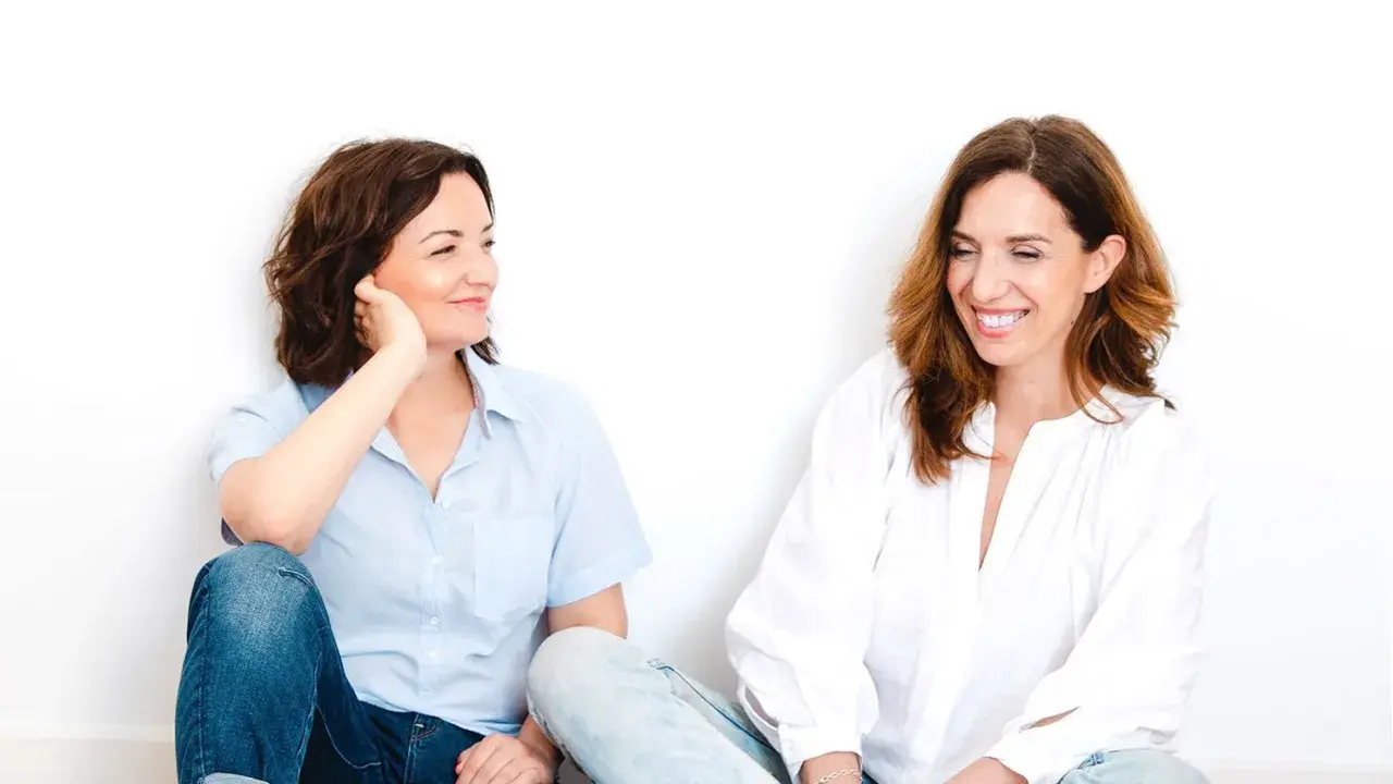 Dos mujeres sentadas, una con camisa azul y jeans, la otra con blusa blanca. Parecen compartir un momento laconicum, de pocas palabras pero profunda conexión.