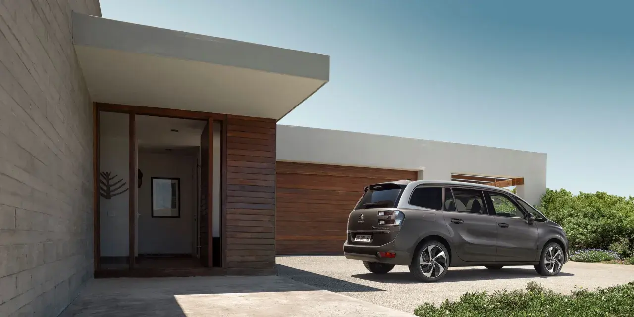 Szary minivan Citroën C4 Picasso zaparkowany przed nowoczesnym domem z drewnianą elewacją.