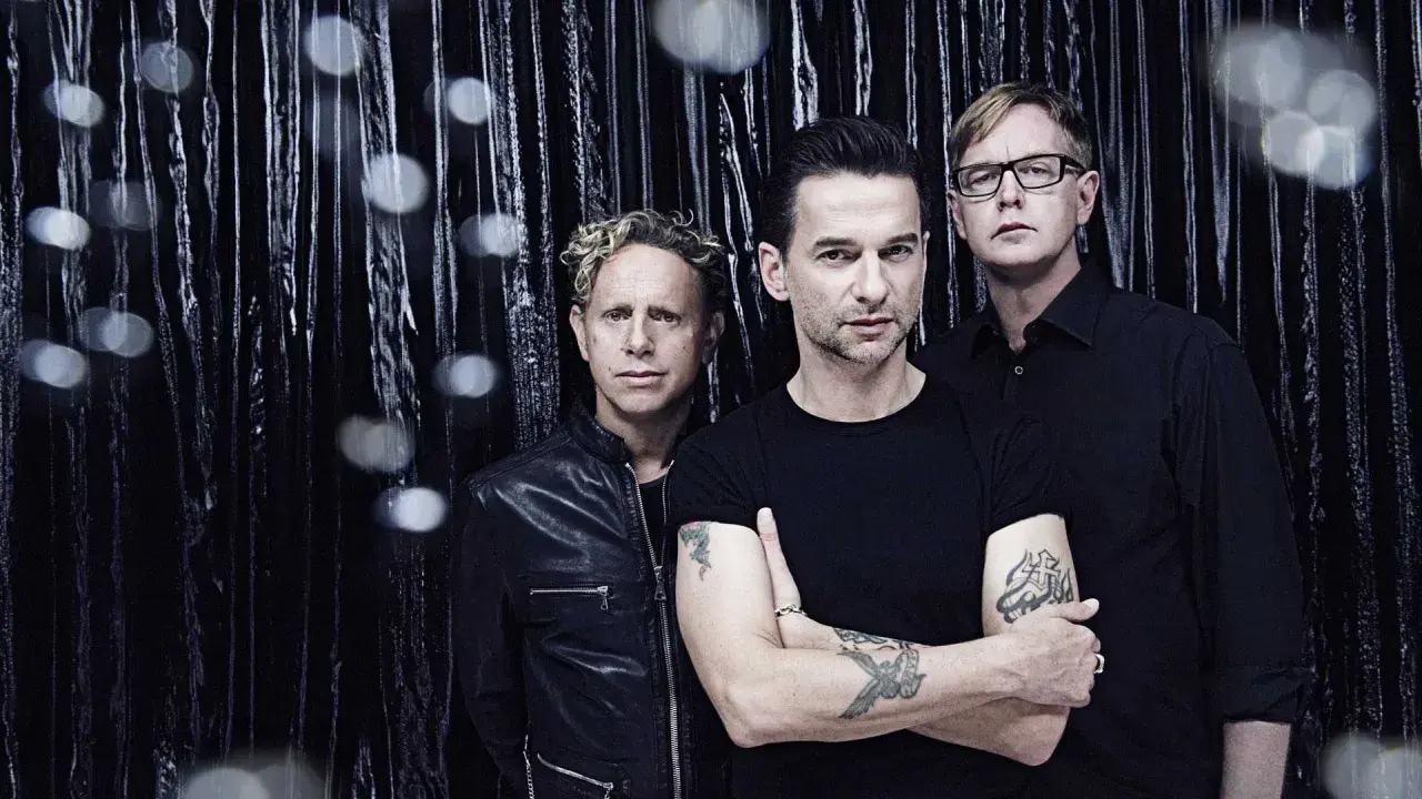 Skład Depeche Mode: Martin Gore, Dave Gahan i Andy Fletcher pozują na tle błyszczącej, czarnej kurtyny.
