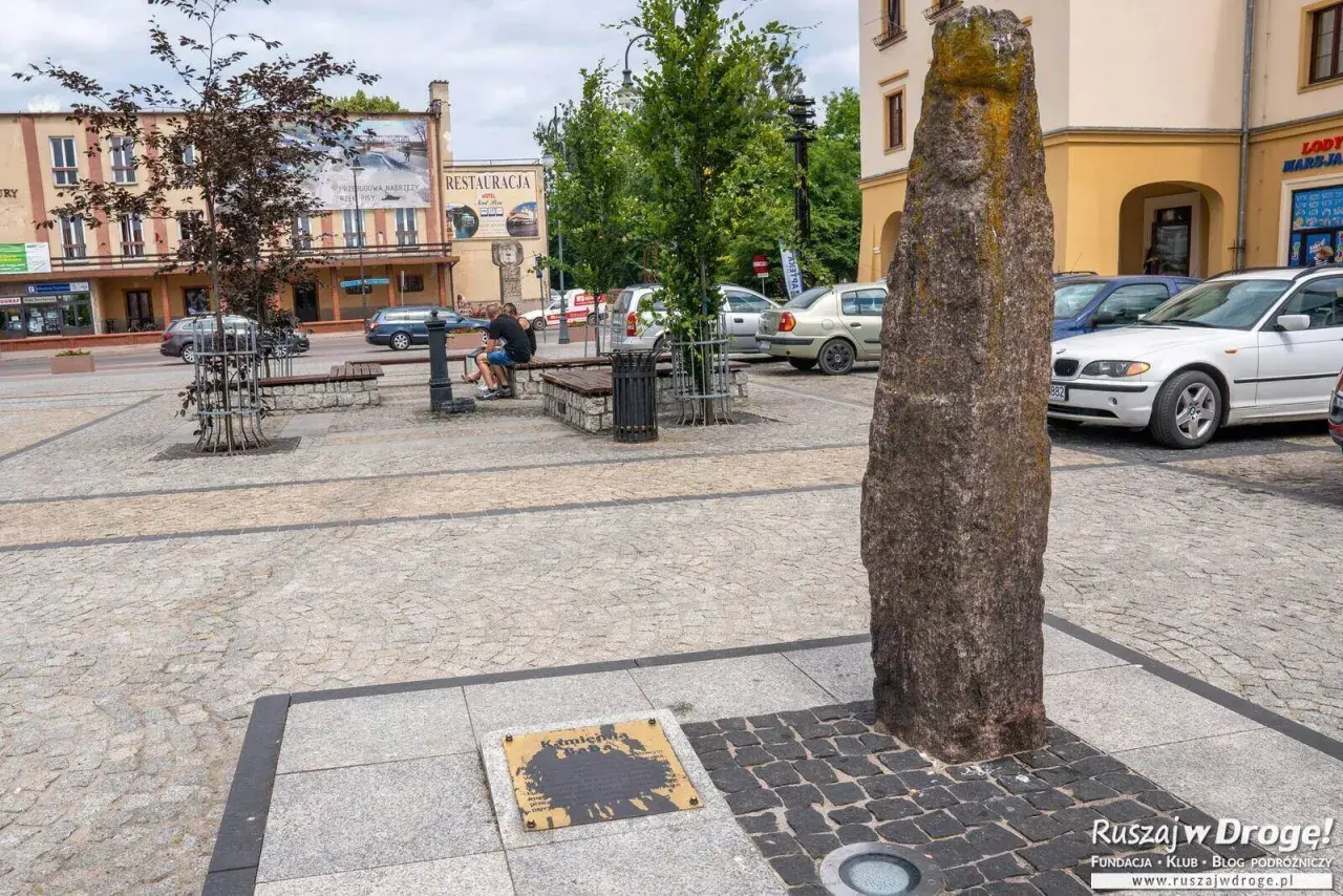 Kamienny pomnik i tablica z napisem "Kamienna" to ciekawe atrakcje na rynku. Obok ławki i drzewa, w tle budynki i samochody.