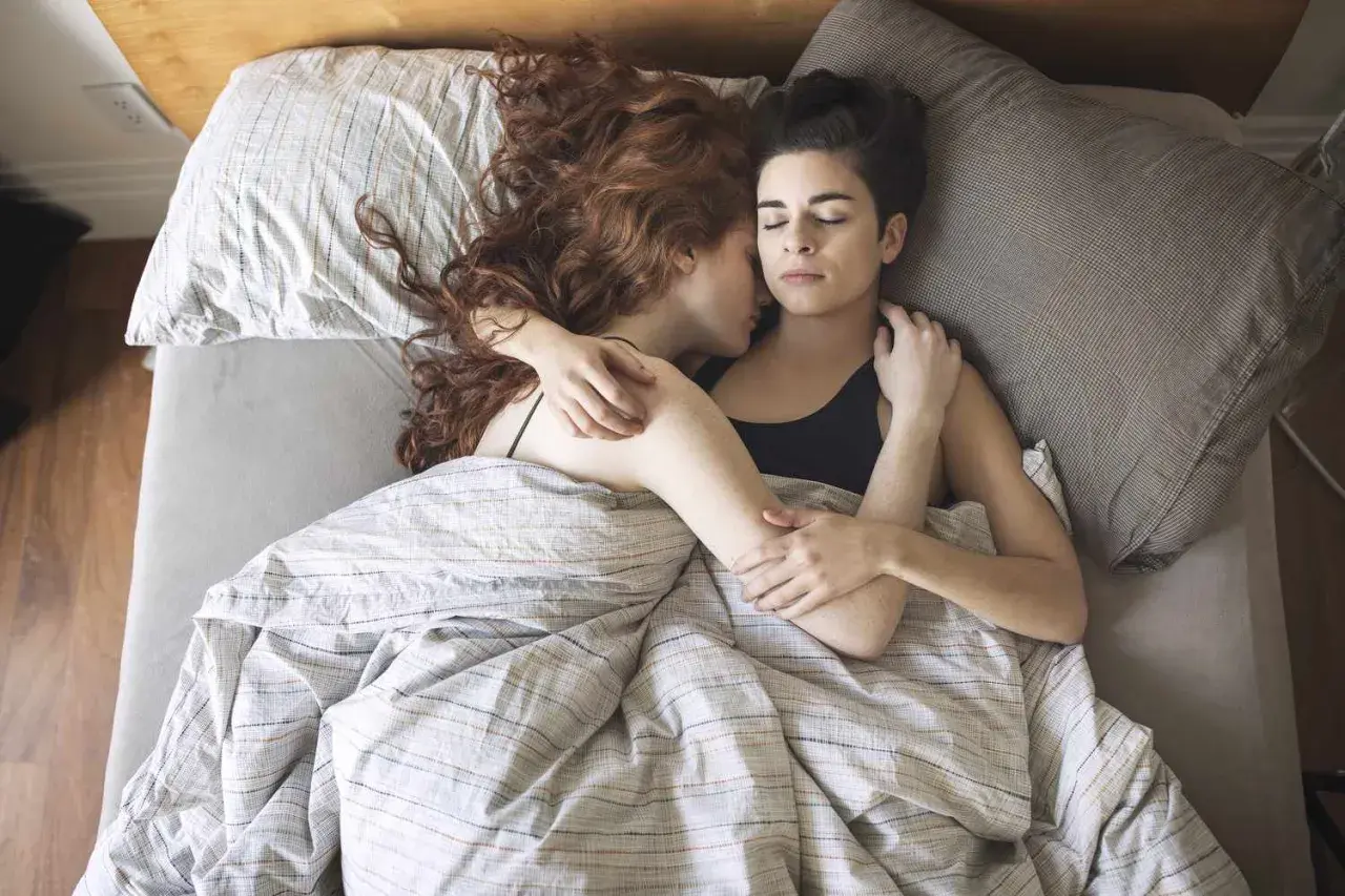 Two women sleeping together, embracing warmly under a striped duvet.