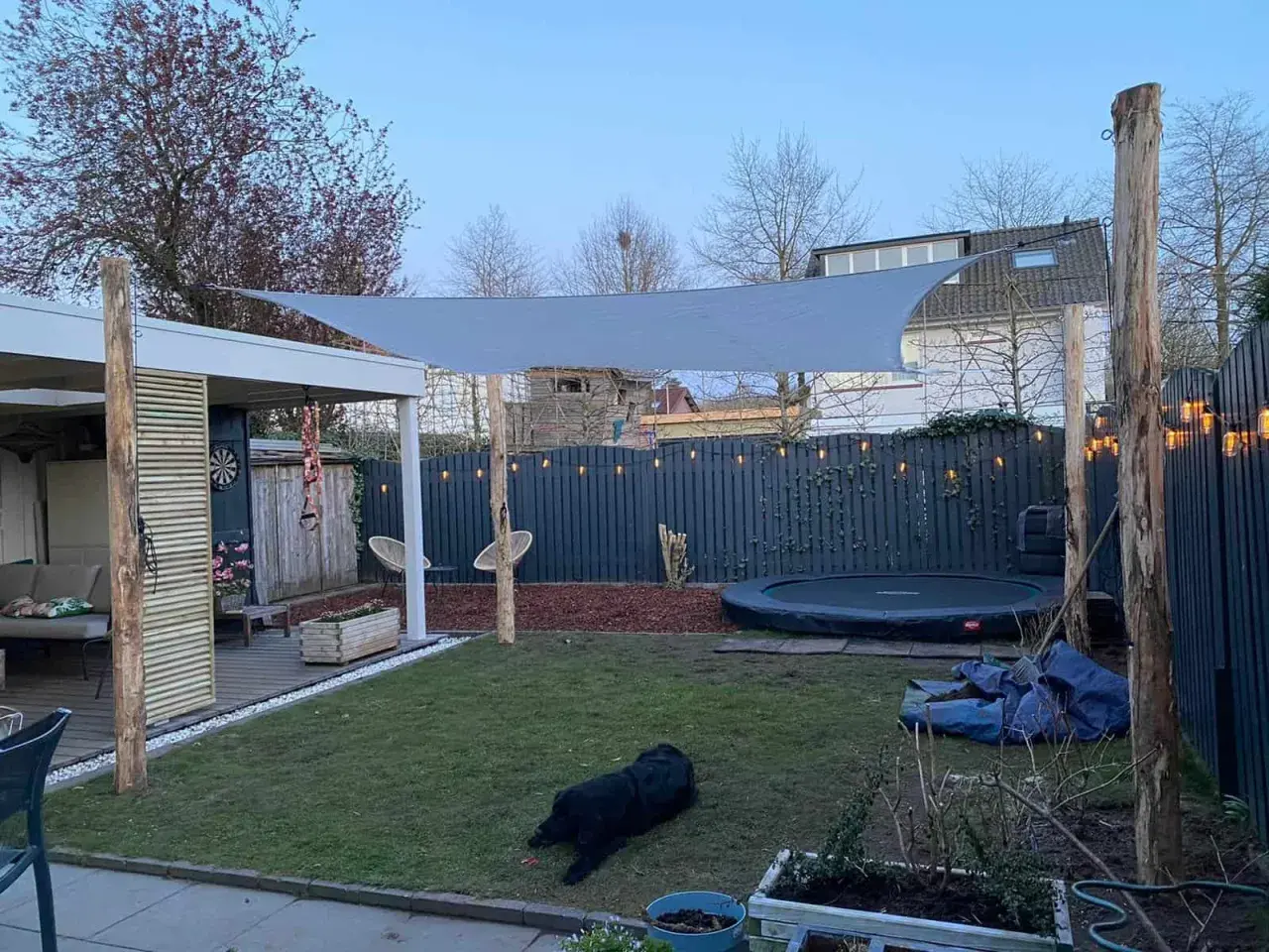 Sonnensegel befestigen Ideen: Ein graues Sonnensegel spannt sich über einen Garten mit Trampolin und Sitzbereich. Holzpfosten dienen als Befestigungspunkte.