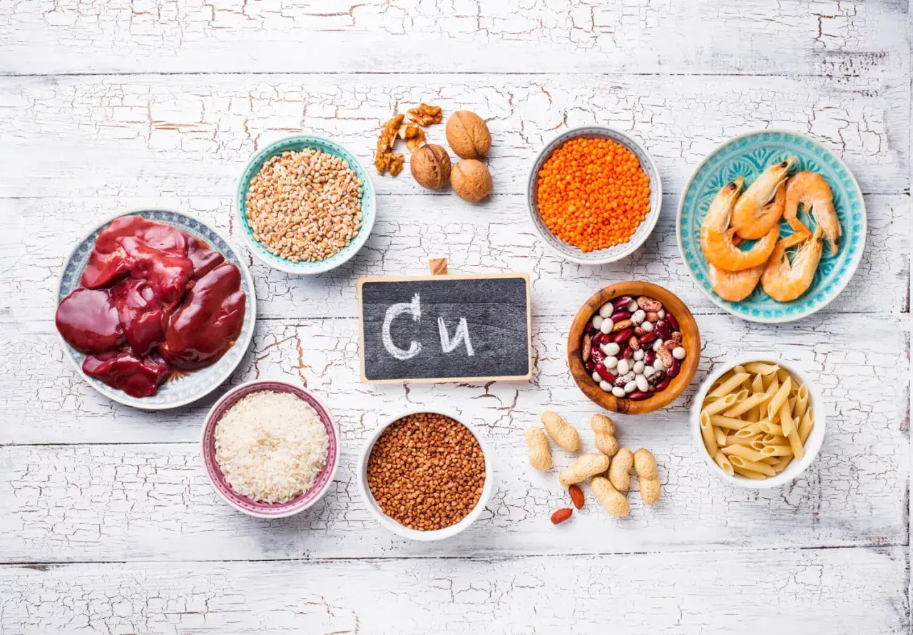 Na tabliczce napisano "Cu". Wokół niej są produkty bogate w miedź: wątróbka, soczewica, fasola, orzechy, krewetki, ryż, kasza gryczana, pszenica, makaron i orzeszki ziemne. Czy miedź rdzewieje? Nie, ale jest niezbędna dla zdrowia.