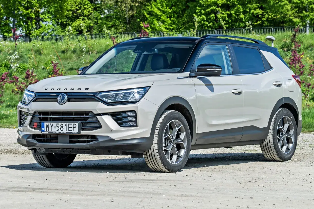 Jasnoszary SsangYong Korando z czarnym dachem stoi na drodze gruntowej, otoczony zielenią.