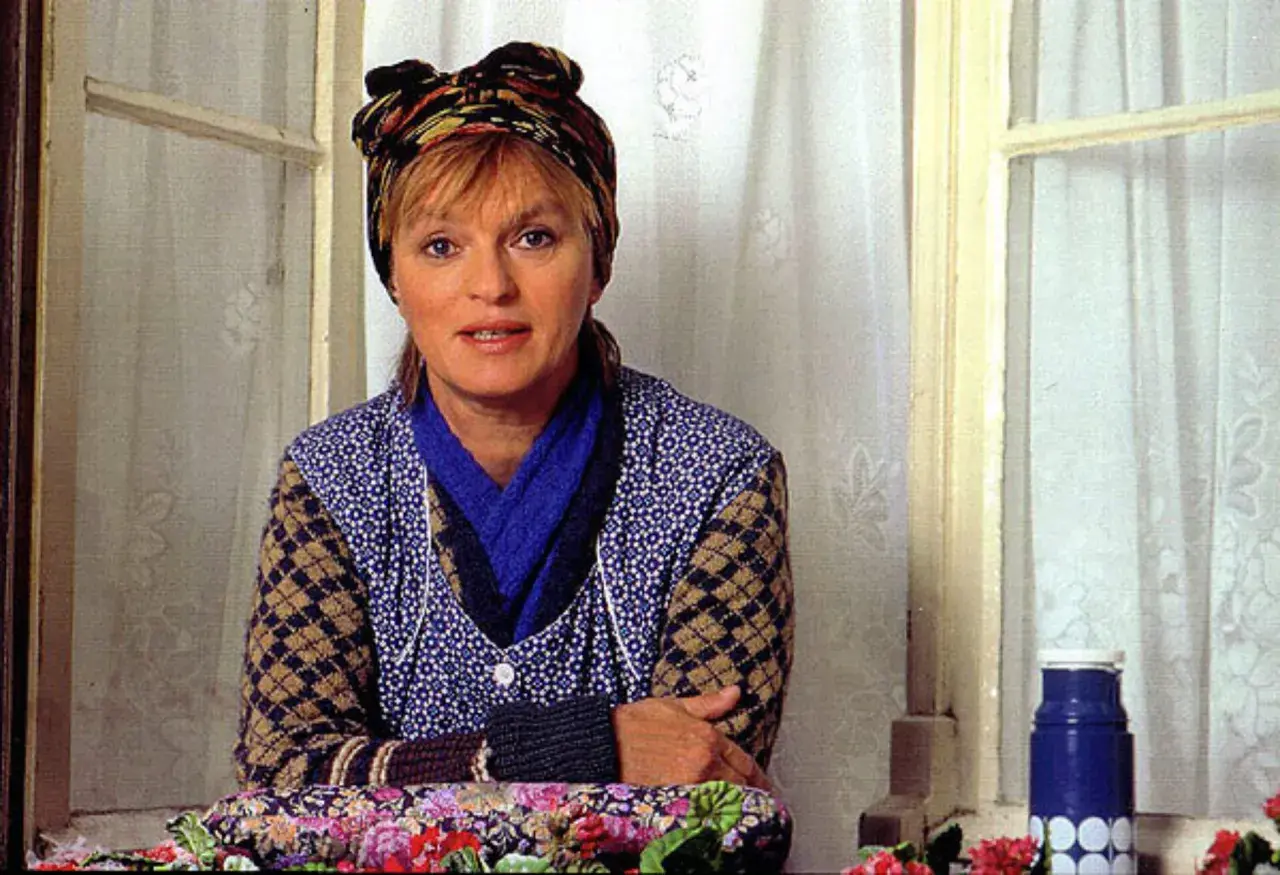 Else Stratmann sitzt am Fenster, eine bunte Kopftuch ziert ihr Haar. Sie trägt eine blaue Bluse mit weißen Punkten und eine Strickjacke.