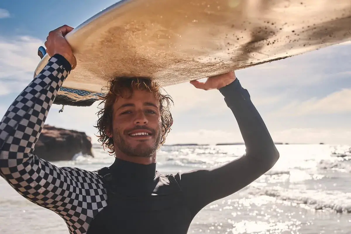 Lächelnder Surfer mit lockiger surfer frisur trägt Neoprenanzug und hält Surfbrett über dem Kopf.