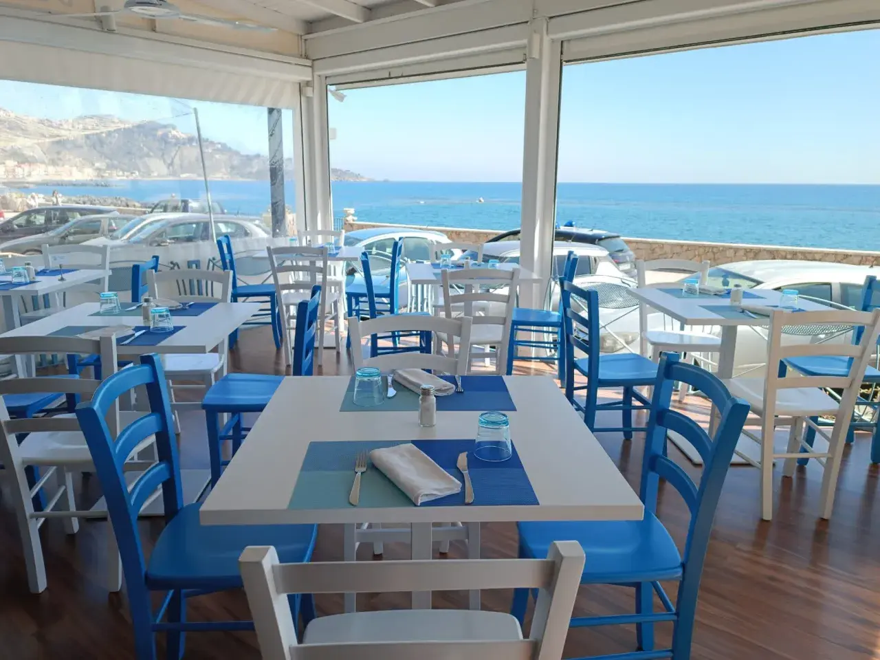 Tavoli apparecchiati con vista mare, perfetti per un apericena a Giardini Naxos. Sedie bianche e blu, un'atmosfera rilassante.