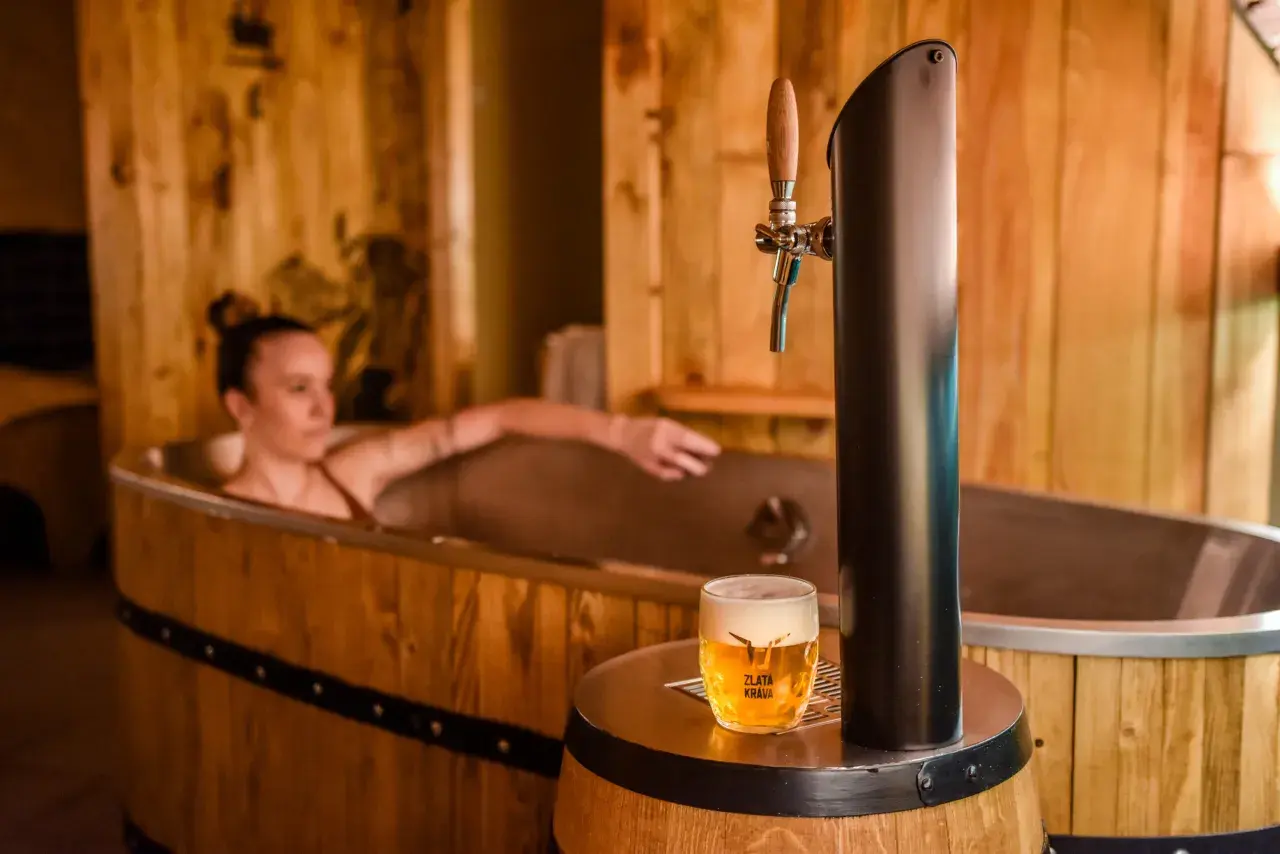 Entspannende Frau in einem Bierfass-Bad, daneben ein frisch gezapftes Pils. Ein einzigartiges Bier-Spa-Erlebnis in Pilsen.