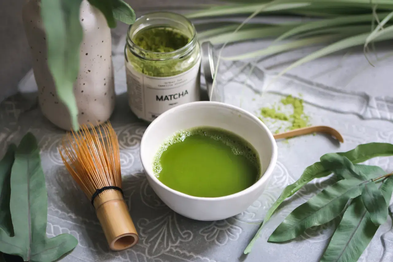 Matcha-Pulver in einem Glas, eine Schale mit zubereitetem Matcha und ein Bambusbesen. Die Matcha-Wirkung auf den Darm wird oft positiv beschrieben.