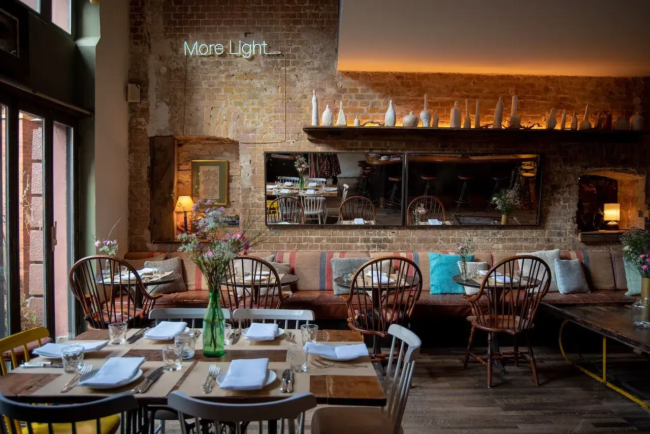 Gemütliches Restaurant mit Ziegelwänden und Tischen, gedeckt für Gäste. Eine leuchtende Neon-Schrift "More Light" hängt über dem Sitzbereich.