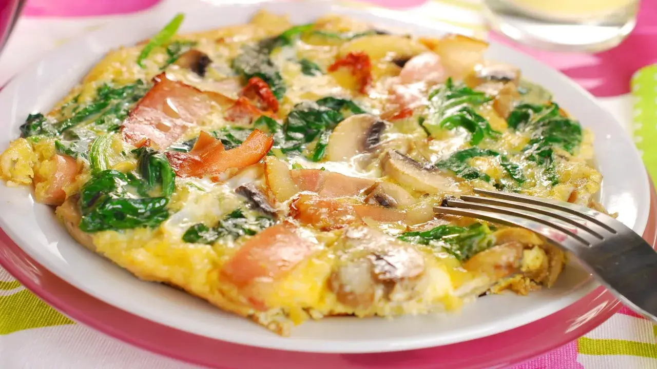 Omlet z szpinakiem, pieczarkami i bekonem na białym talerzu, widelcem.