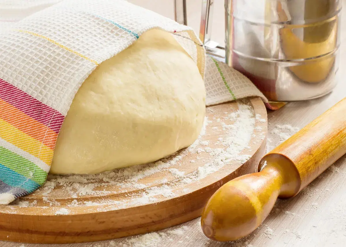 Masa de pan cubierta con un paño a rayas, sobre una tabla de madera con harina esparcida. Rodillo y tamizador de harina, ingredientes para hacer pan.