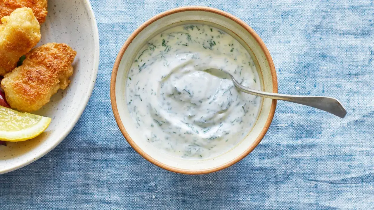 En skål med krämig kall sås till fisk, med dill och en sked. Bredvid syns friterad fisk och citronklyftor.