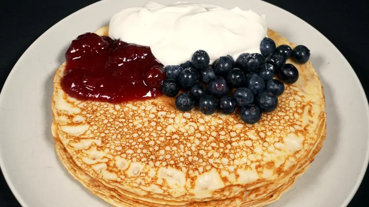 En hög med glutenfria pannkakor toppas med sylt, blåbär och grädde.