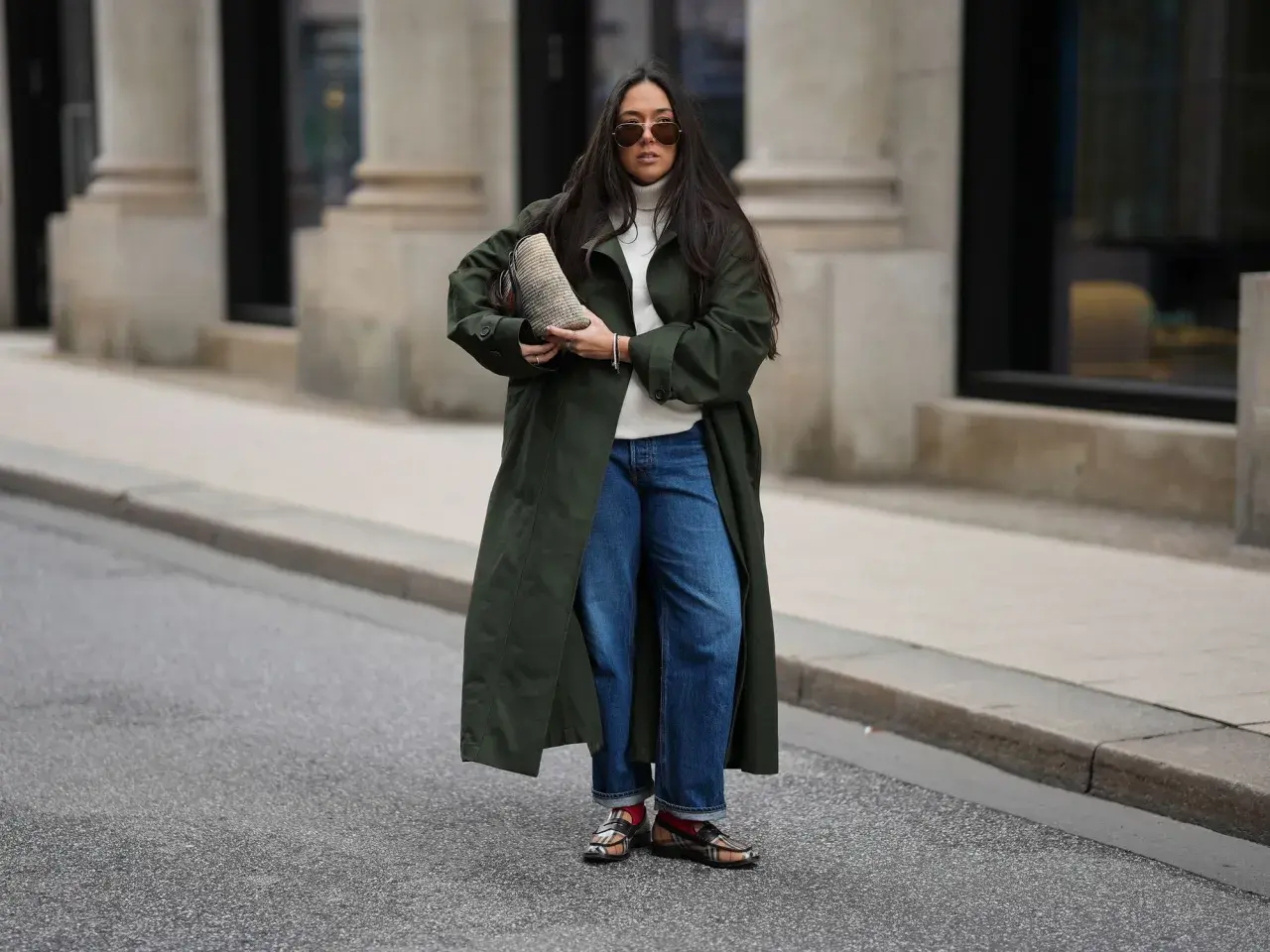 Stylische Frau in dunkelgrünem Mantel und Barrel Jeans, die sie lässig kombiniert. Sie trägt eine weiße Rollkragenpullover und auffällige Schuhe.