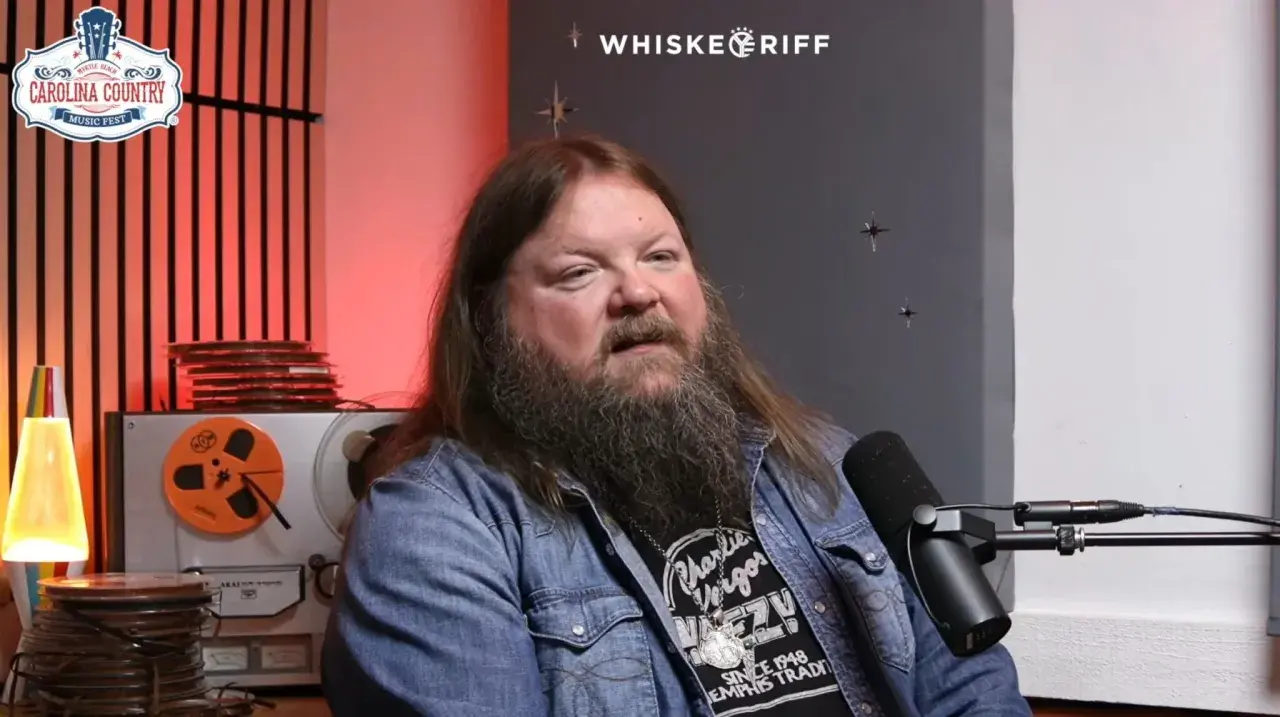 A man with a long beard and hair, wearing a denim jacket, speaks into a microphone. He's discussing Channing Wilson songs, possibly at a music festival.