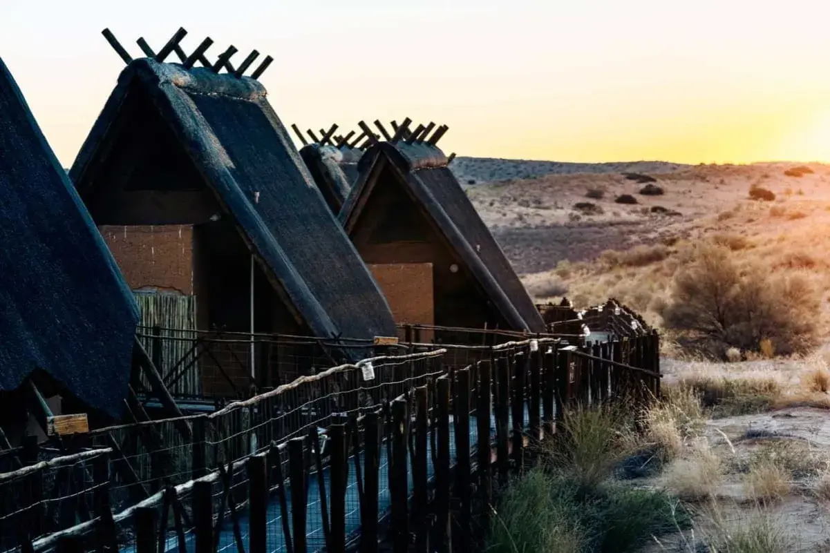 Hütten mit Strohdächern im Kgalagadi Transfrontier Park bei Sonnenuntergang. Ein Holzsteg führt zu den Unterkünften.
