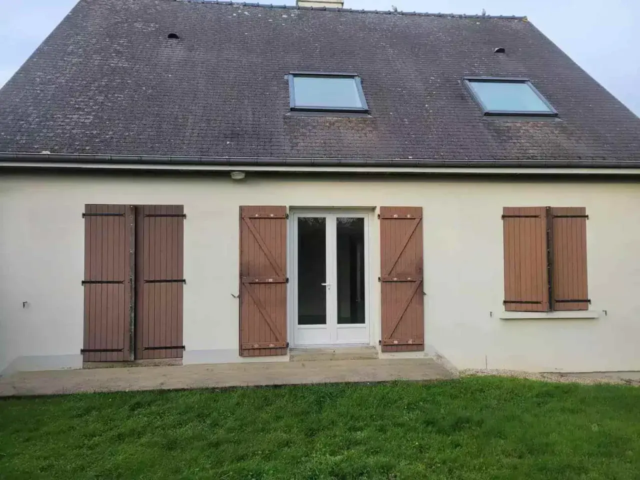 Façade d'une maison année 80, caractéristiques : toit en ardoise, deux fenêtres de toit, porte-fenêtre blanche et volets en bois marron.
