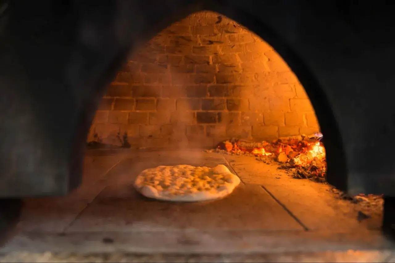 Wnętrze pieca do pizzy z palącym się drewnem i surowym ciastem na kamiennej podłodze. Dym unosi się wokół.