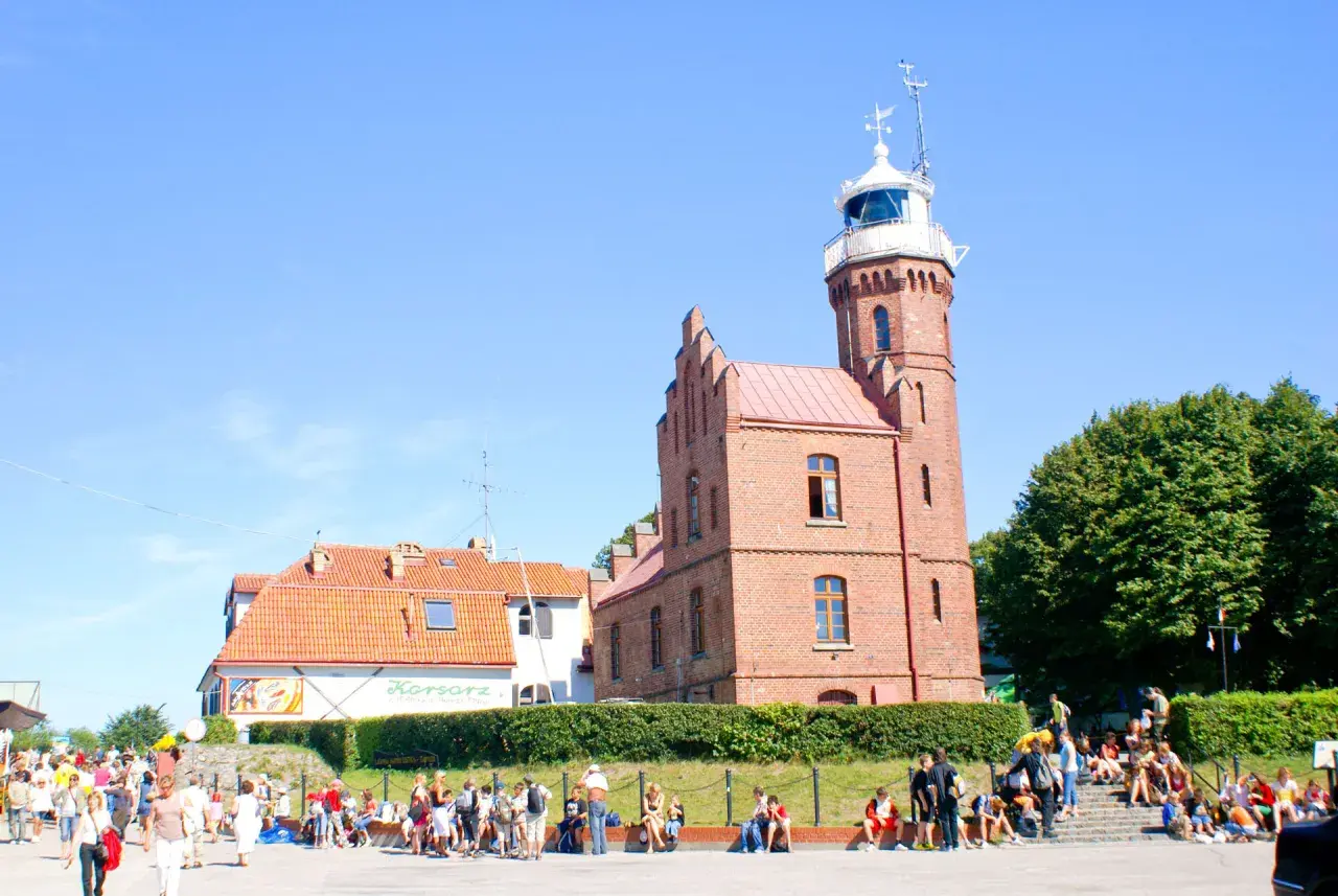 Urokliwa latarnia w Ustce, otoczona zielenią i tłumem turystów.