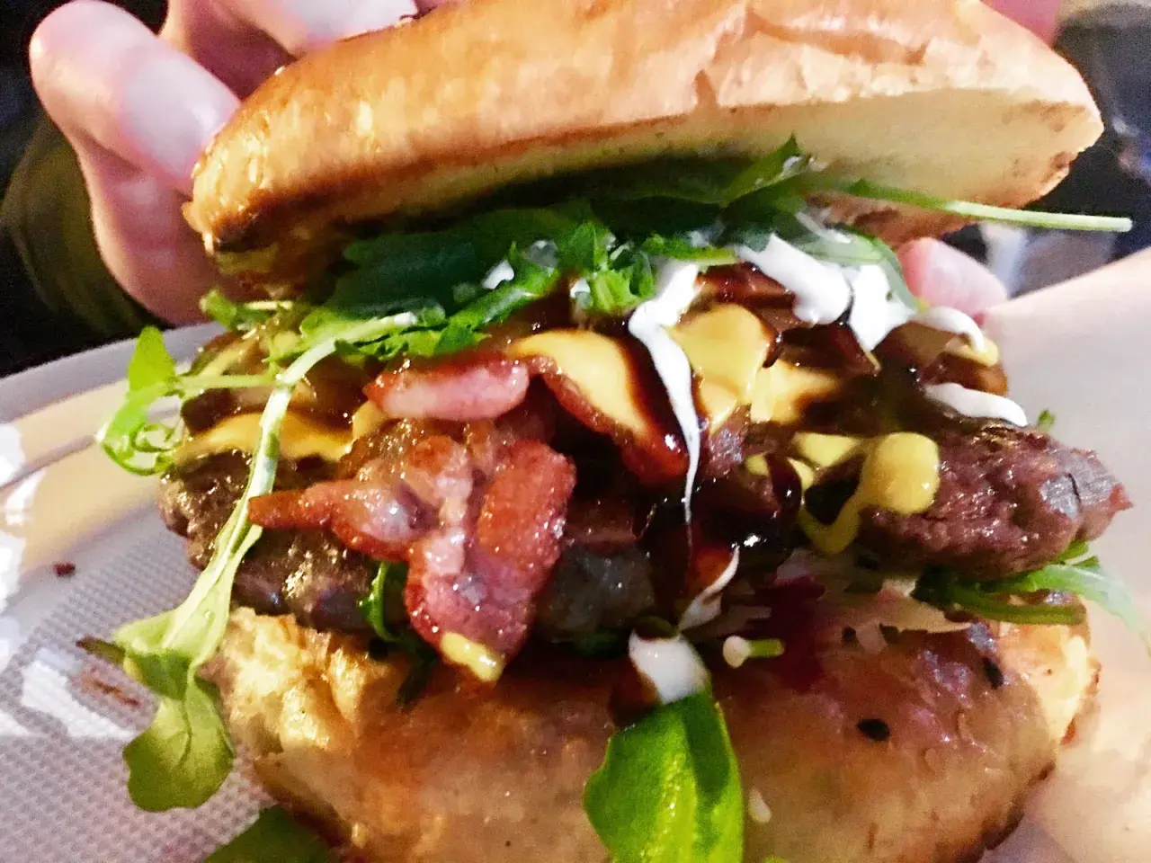 Soczysty burger z bekonem, serem, rukolą i sosami, trzymany w dłoni.