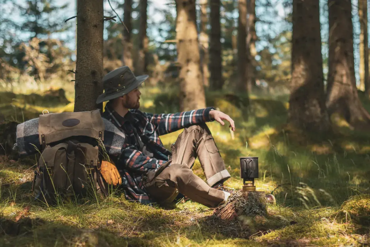 Man med hatt och rutig skjorta vilar vid ett träd under en vandring i Småland. Ryggsäck och stormkök redo för äventyr.