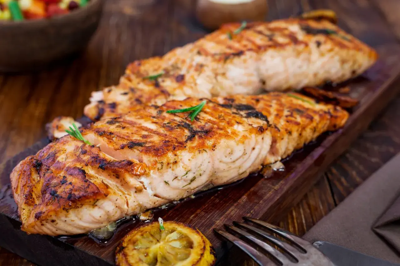 Två saftiga filéer av grillad fisk, perfekt kryddad med örter, serveras på en träbricka med citron och en gaffel.