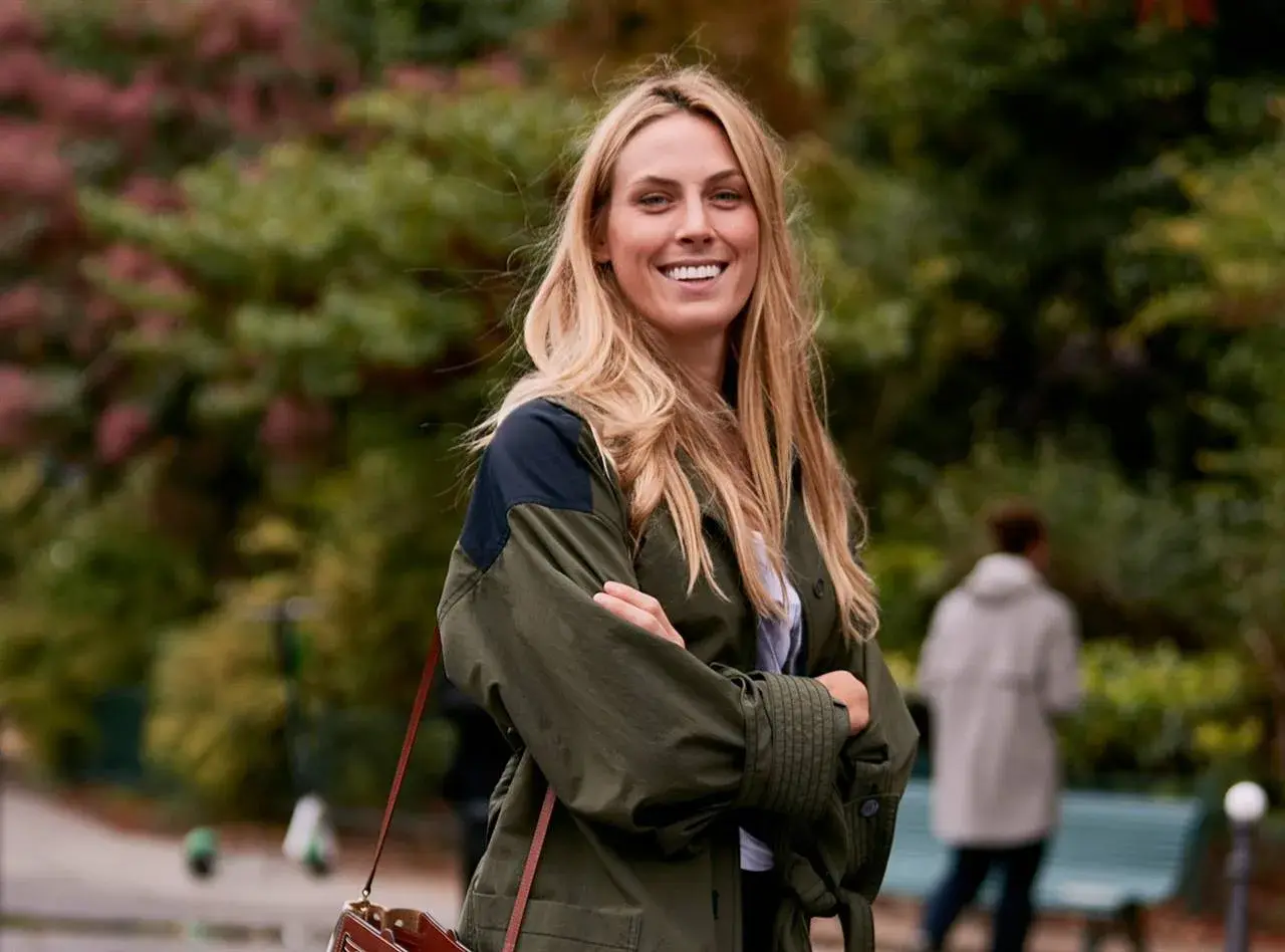 Mujer sonriente con un corte de pelo a capas, luciendo una chaqueta verde y un bolso de cuero.