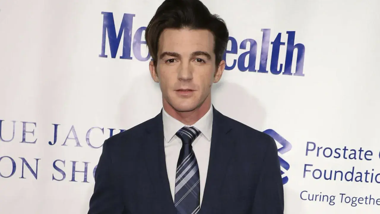 Drake Bell w garniturze, znany z filmów, seriali i programów, pozuje na tle wydarzenia charytatywnego.