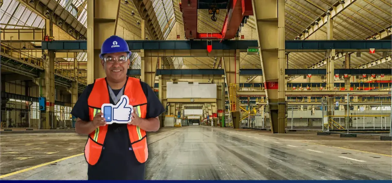 Hombre con casco y chaleco naranja sonríe en una nave industrial, sosteniendo un pulgar hacia arriba. Sus sistemas de limpieza son eficientes.